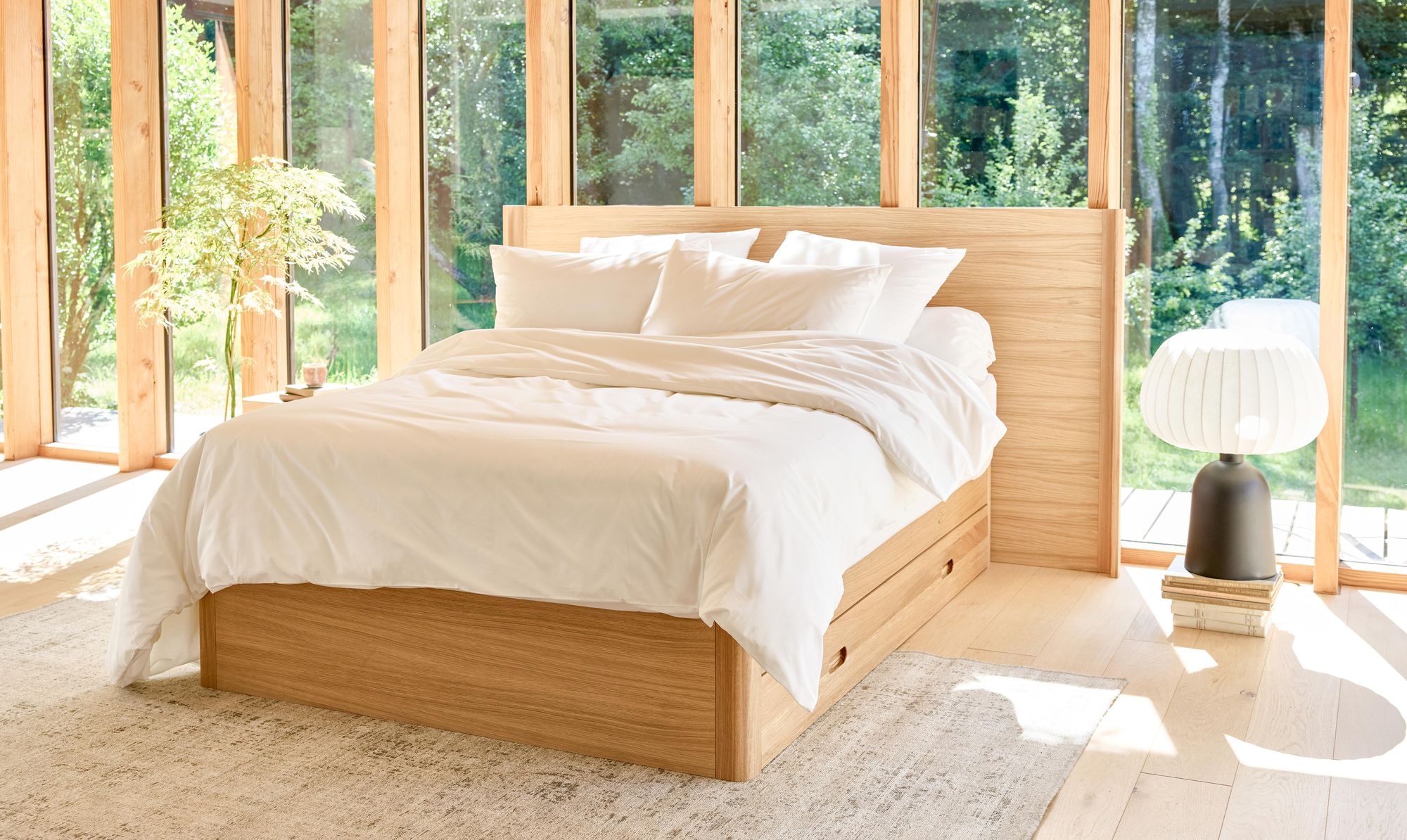 Chambre lumineuse avec lit en chêne à tiroirs Kipli, draps blancs et lumière naturelle traversant de grandes baies vitrées.