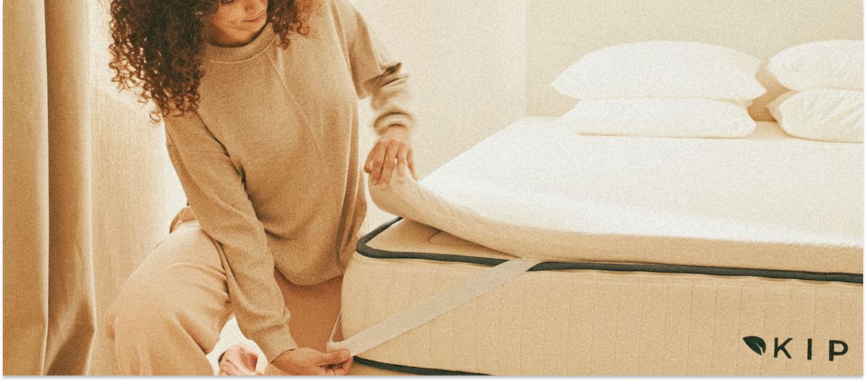 Femme installant un surmatelas sur un matelas en latex naturel Kipli dans une chambre lumineuse et minimaliste.