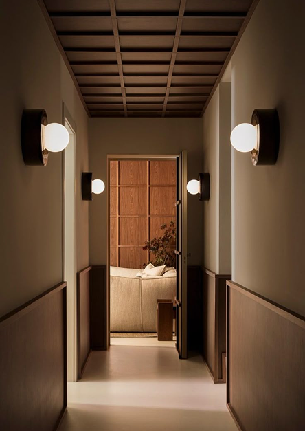 Couloir minimaliste éclairé par des appliques murales modernes, menant à un salon chaleureux en bois et canapé beige, illustrant une décoration intérieure élégante et apaisante.