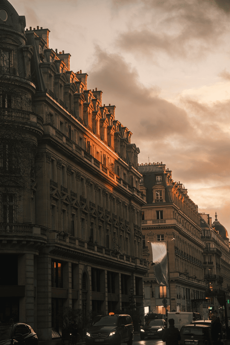 Façades haussmanniennes à Paris au coucher du soleil, avec lumière dorée sur les toits et circulation en bas des immeubles.